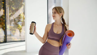 Woman in yoga clothes with mat is standing indoors with drink in hand. Making a break