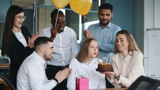 Young woman is working in office then getting birthday cake from colleagues making wish blowing candle enjoying surprise having fun with creative team