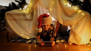 Happy little boy in Santa hat lying in DIY child tent celebrating Christmas using smartphone, having video call. Christmas at home.
