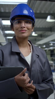 Young woman engineer wearing a blue helmet and work coat posing at a factory holding a tablet, portrait shot. Female professionals in industry concept.