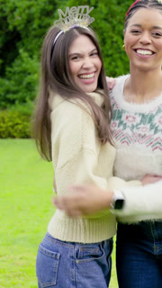 Vertical video: Posing after camera cue, two women embracing on lawn wearing headbands and sweaters