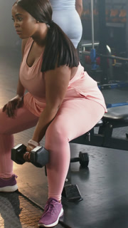 Vertical video: Gripping seated woman in pink curling hex dumbbell on gym bench for arms