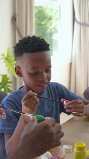 Vertical video: Handing paint jars, adult hands guiding boy painting Easter egg at sunlit table