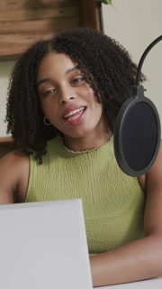 Vertical video: Recording host in green top speaking into pop-filter mic at home checking laptop