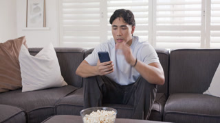 Asian man 20s wearing tee reading message on blue phone on sofa, grabbing popcorn, fist-pumping