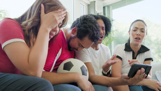 Friends in jerseys leaning on couch, watching key play on phone, bearded man holding ball, reacting