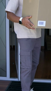 Vertical video: Stepping on mat, adult man wearing beanie carrying box into home, inspecting label