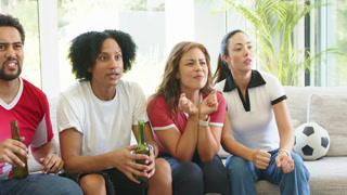 Diverse friends on couch watching soccer match, reacting to tense play, holding beers, soccer ball