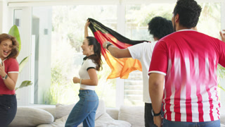 Friends in jerseys reacting to match lifting black-red-gold scarf and raising soccer ball at home
