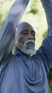 Vertical video: Breathing senior man bringing hands together in garden, meditating in gray pullover