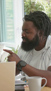 African American man sitting at table near window, gesturing and pointing at laptop with mug
