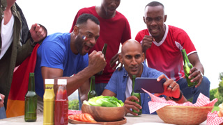 Diverse male friends leaning over picnic table watching smartphone clip, cheering with beer bottles