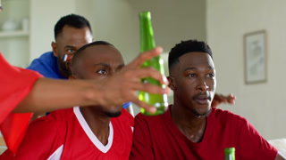 Three male friends in red and blue tees reacting to offscreen game, pointing, raising green bottle