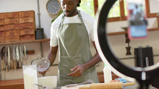 African man in apron filming on phone, sieving flour, cracking egg in kitchen for lesson