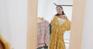 Choosing between outfits, asian woman holding dress and sweater in front of mirror, at home