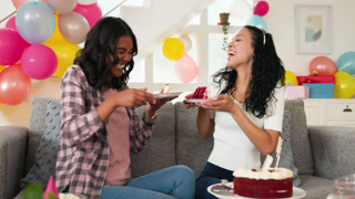 Diverse female friends enjoying cake and popcorn on gray sofa in living room, with balloons