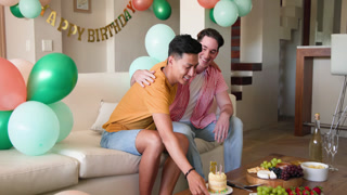Celebrating birthday, gay couple enjoying cake and laughter on living room sofa at home