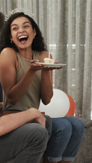 Vertical video: Diverse couple on sofa sharing birthday offering candle cake blowing and laughing