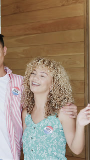 Vertical video: Couple embracing on porch raising, waving US flag with VOTE pins celebrating voting