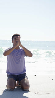 Vertical video: Sunlight guiding African American male stretching into prayer pose at beach