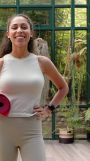 Vertical video: Woman shifting onto leg, smiling into laughter holding mat in greenhouse for yoga