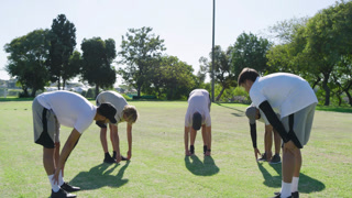 Stretching diverse male teammates on grassy field under clear sky, with neon cone, lamppost
