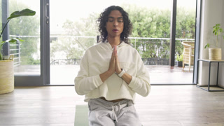Closing eyes and pressing palms, non-binary adult meditating for calm in living room on yoga mat