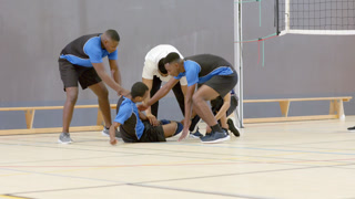 Asian man collapsing onto gym floor near net, teammates and coach rushing in, examining injured leg