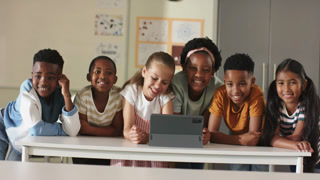 Six diverse schoolchildren leaning on table in classroom, peering at tablet on stand and smiling