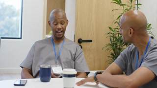 Two male coworkers in scrubs, right-leaning and gesturing, talking at break table with smartphone