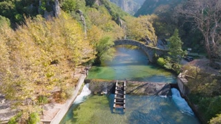 Aerial drone flight over the Voidomatis River, approaching the historic Klidonia stone bridge in Zagori, Greece. Shot reveals the waterfalls, autumn trees, and gorge