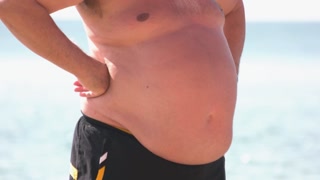 Aged abased man with belly fat standing on the beach. Unhealthy lifestyle concept.