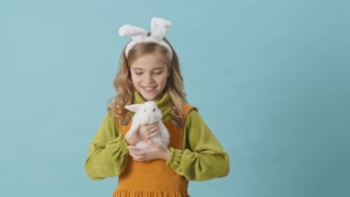 Cheerful young girl with blond hair and bunny ears is holding a rabbit in her hands and smiling isolated over blue background