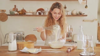 A positive pleased young woman is stirring ingredients in a bowl at a cozy kitchen