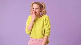 A cute blonde girl heard a funny joke and shyly laughs at her, covering her mouth with her hand in an isolated studio on a purple background