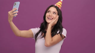 Happy birthday girl with a cap on her head accepts online congratulations using her mobile standing isolated over violet background in the studio