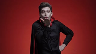 Vampire man with blood and fangs in black halloween costume blowing a kiss isolated over red wall