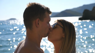 Close up of young couple kissing near seashore on sunny day. Beautiful pair spending time together on summer vacation. Sun light reflection at the sea water surface at background. Concept of loving.