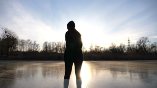 Girl in figure skates is skating on ice surface at sunset time. Young woman training alone at frozen river or lake. Lady improves skills and have active leisure at winter cold weather. Slow motion.