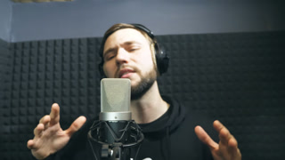 Young man emotionally recording new melody or song. Male singer in headphones singing song into the microphone at sound studio. Working of creative musician. Show business concept. Slow motion.