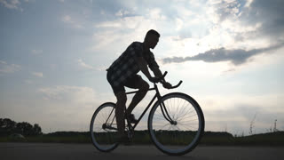 Silhouette of young man riding at vintage bicycle with beautiful sunset sky at background. Sporty guy cycling in the country road. Male cyclist riding fixed gear bike. Healthy active lifestyle Closeup.