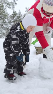 Grandmother and child building a snowman in snowy forest. Cozy winter clothing, family love, healthy outdoor lifestyle and joyful holiday