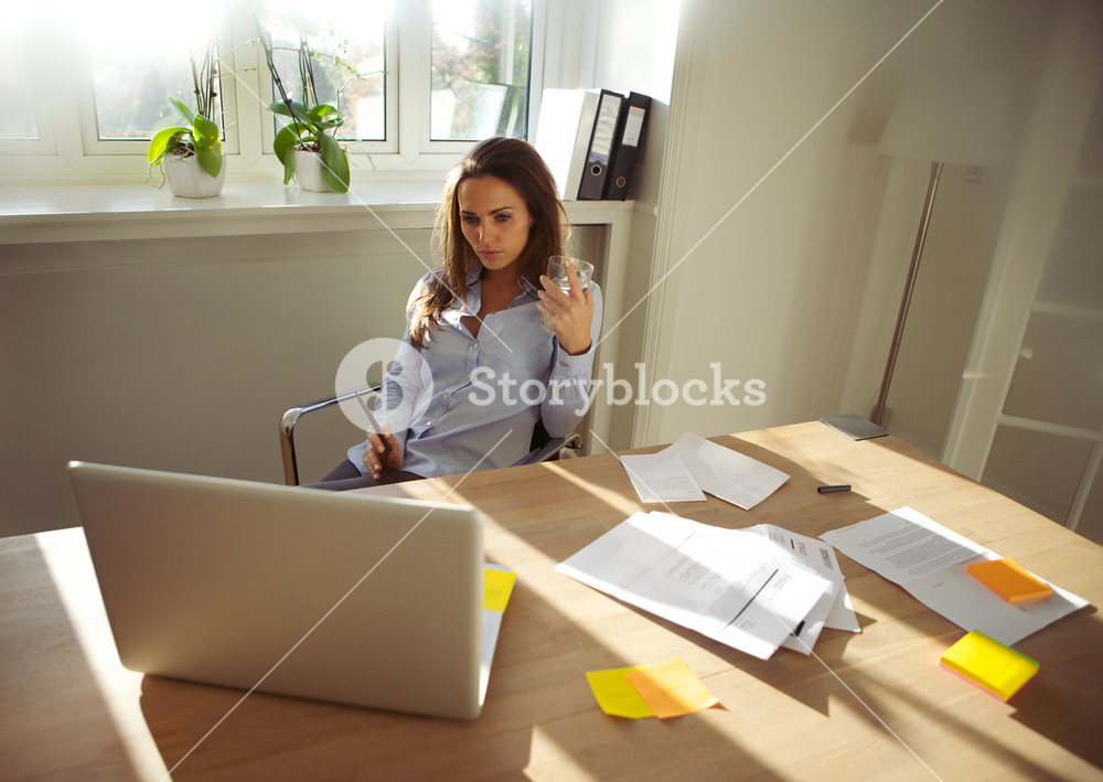 Young businesswoman holding a glass of water, focused on laptop for new project. Woman working on business plan from home office.